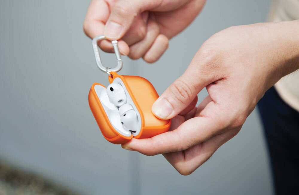 Osoba trzyma pomarańczowe etui na słuchawki, w którym znajdują się białe słuchawki bezprzewodowe. Etui jest wyposażone w metalowy karabińczyk.