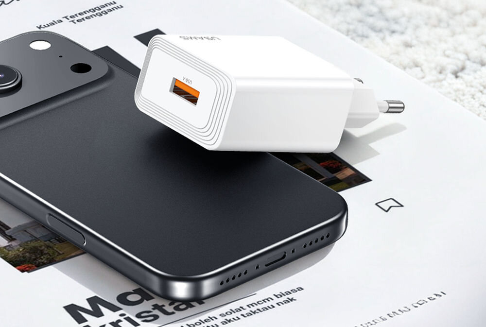 Na obrazie widoczna jest biała ładowarka sieciowa z logo \'USAMS\' oraz czarny smartfon leżący na papierze z tekstem. Ładowarka ma pomarańczowy port USB i wtyczkę do gniazdka elektrycznego.