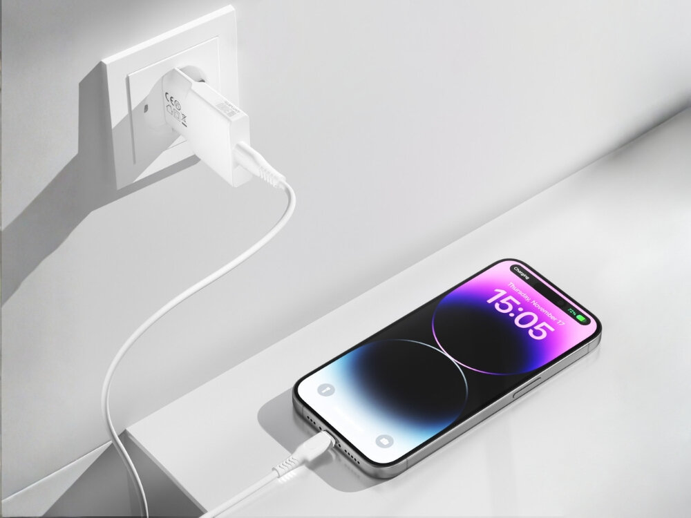 Biała ładowarka sieciowa podłączona do gniazdka, z kablem USB-C łączącym się z telefonem leżącym na powierzchni. Na ekranie telefonu widoczna jest godzina 15:05.
