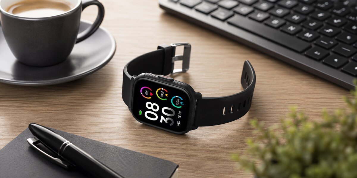 Na biurku leży czarny smartwatch Denver. Obok znajduje się filiżanka kawy, czarny notes oraz klawiatura.