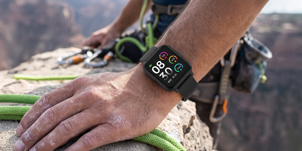 Wspinacz odpoczywa na skale w górach. Na jego nadgarstku znajduje się czarny smartwatch Denver z włączonym ekranem.
