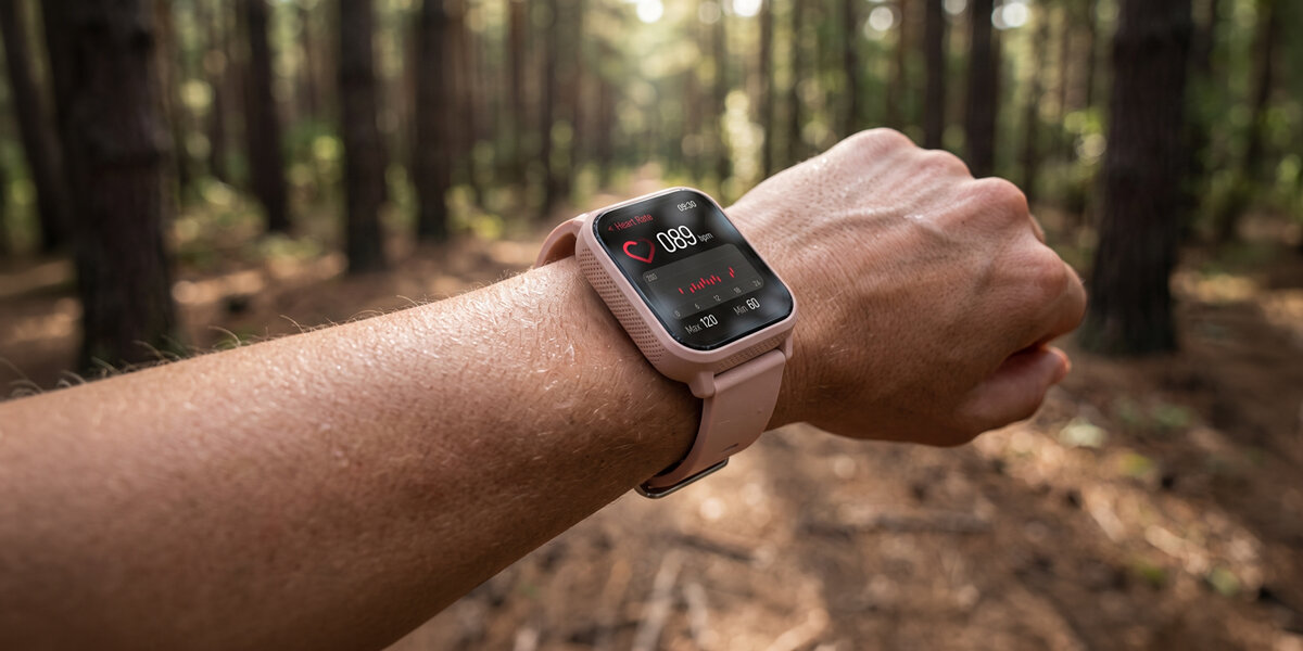 Na ręce kobiety jest różowy smartwatch Denver wyświetlający puls. W tle znajduje się gęsty las.