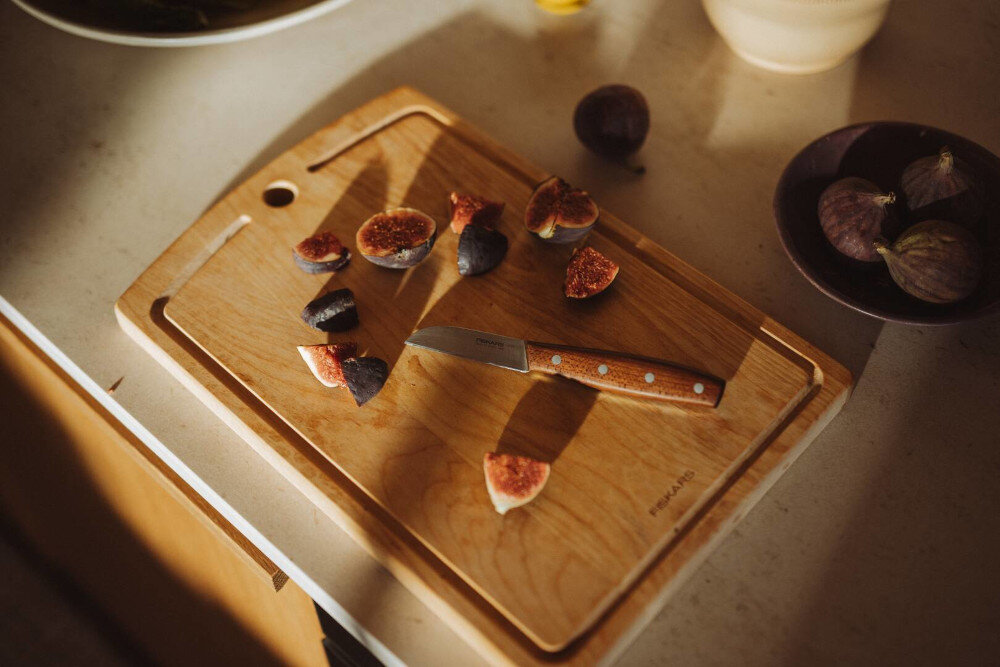 Na drewnianej desce do krojenia leży nóż z drewnianą rączką oraz pokrojone figi. Obok znajduje się miska z całymi figami, a na desce widoczne jest logo \'FISKARS\'.