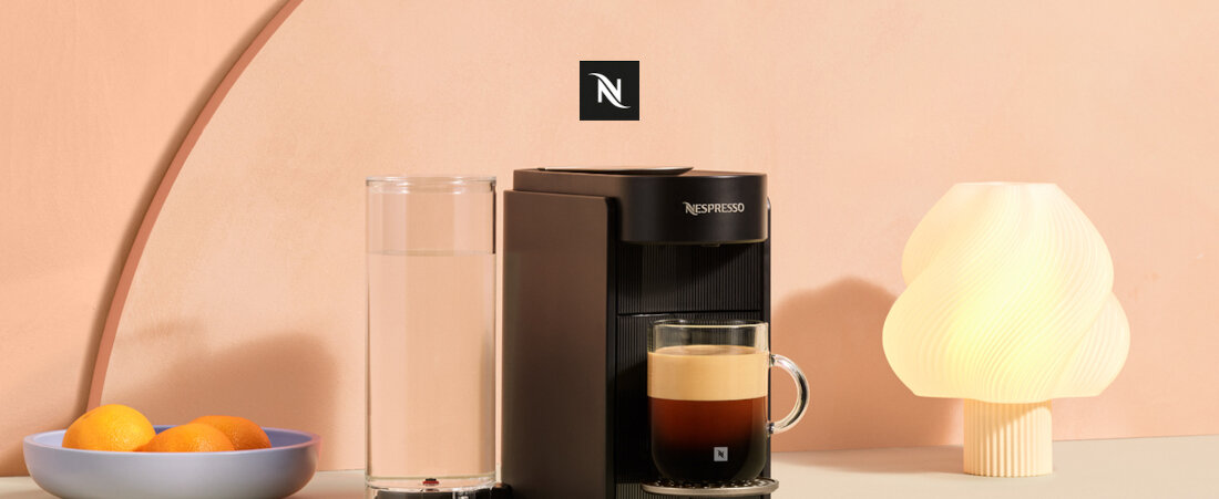 Na obrazie znajduje się czarny ekspres do kawy z filiżanką kawy, obok którego stoi wysoka szklanka z wodą. W tle widoczne jest logo Nespresso oraz lampa o nietypowym kształcie i miska z pomarańczami.
