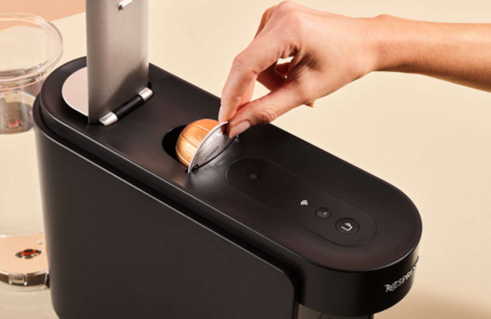 Osoba wkłada kapsułkę do czarnego ekspresu do kawy z widocznym logo \'Nespresso\'.