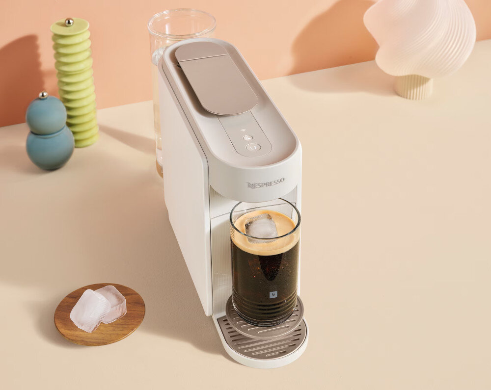 Na blacie stoi biały ekspres do kawy z logo \'Nespresso\', obok którego znajduje się szklanka z kawą i kostkami lodu. W tle widoczne są dekoracyjne przedmioty oraz szklanka z wodą.