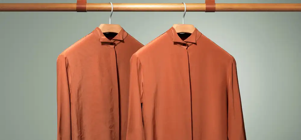 Dwa pomarańczowe koszule wiszą na drewnianych wieszakach na metalowym drążku. Tło jest jednolite i jasne.