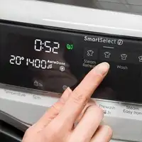 Panel sterowania suszarki z wyświetlaczem pokazującym czas 0:52 i temperaturę 20°C. Widoczna jest ręka naciskająca przycisk \'Steam Refresh\'.