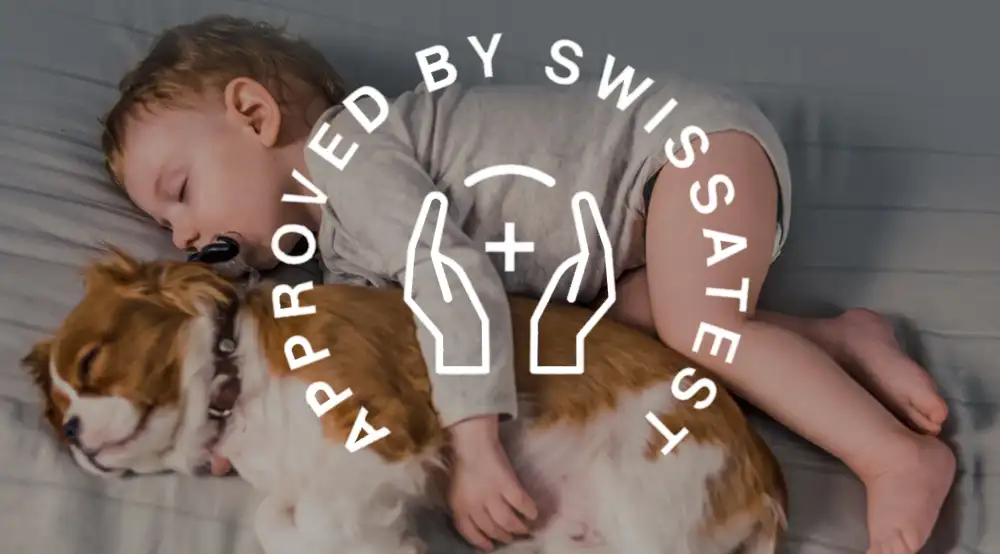 Dziecko śpi obok psa na łóżku. Na obrazie widoczny jest napis \'APPROVED BY SWISSTEST\' oraz symbol dwóch dłoni z plusem.