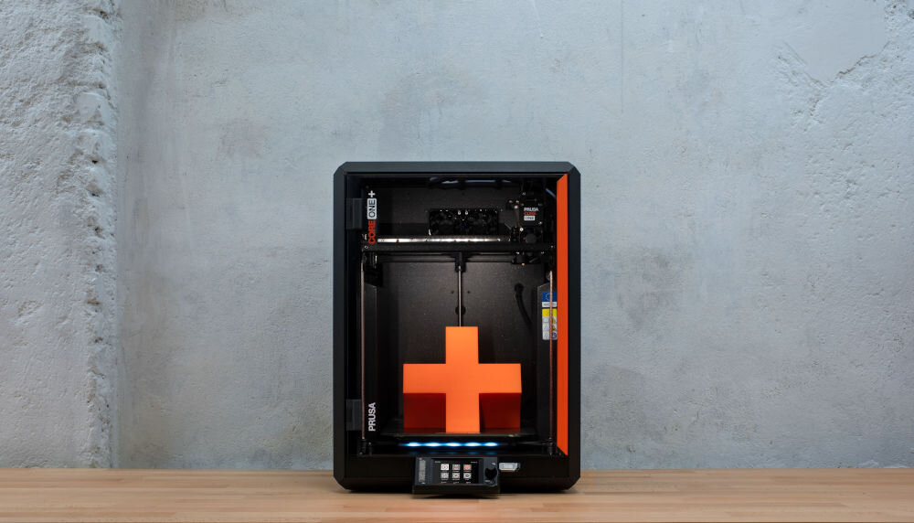 Drukarka 3D PRUSA Core One+ Wi-Fi, LAN (Zamontowana) Widok z przodu na drukarkę 3D z pomarańczowym modelem w środku na jasnym tle wymiary, oszczędność miejsca, pole robocze