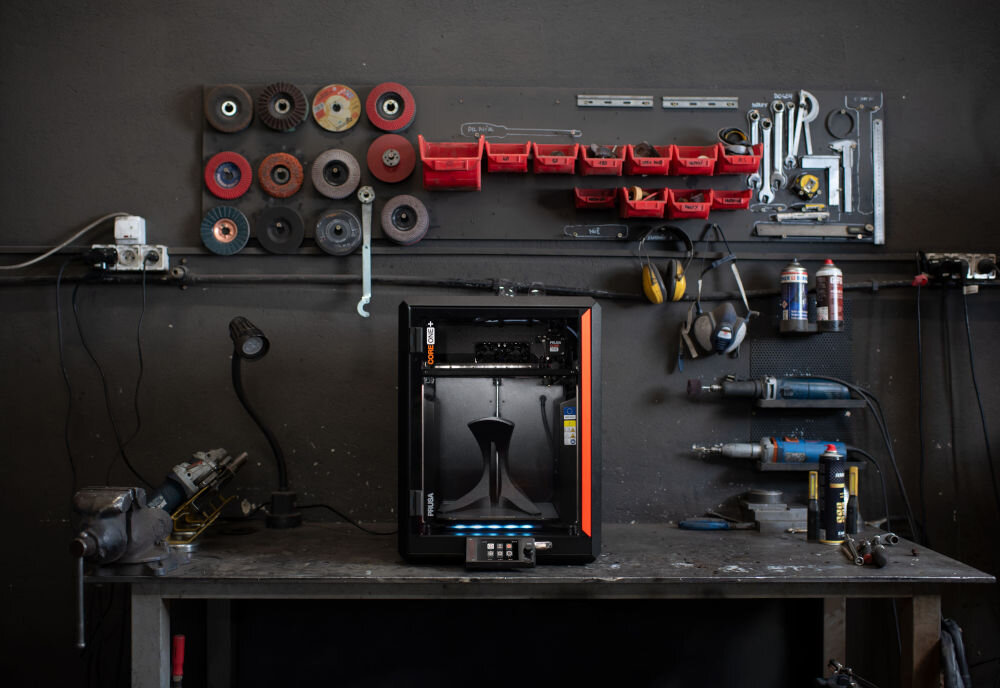 Drukarka 3D PRUSA Core One+ Wi-Fi, LAN + Zestaw do montażu Drukarka 3D na tle ściany z narzędziami zamknięta komora, stabilność cieplna, brak wypaczania