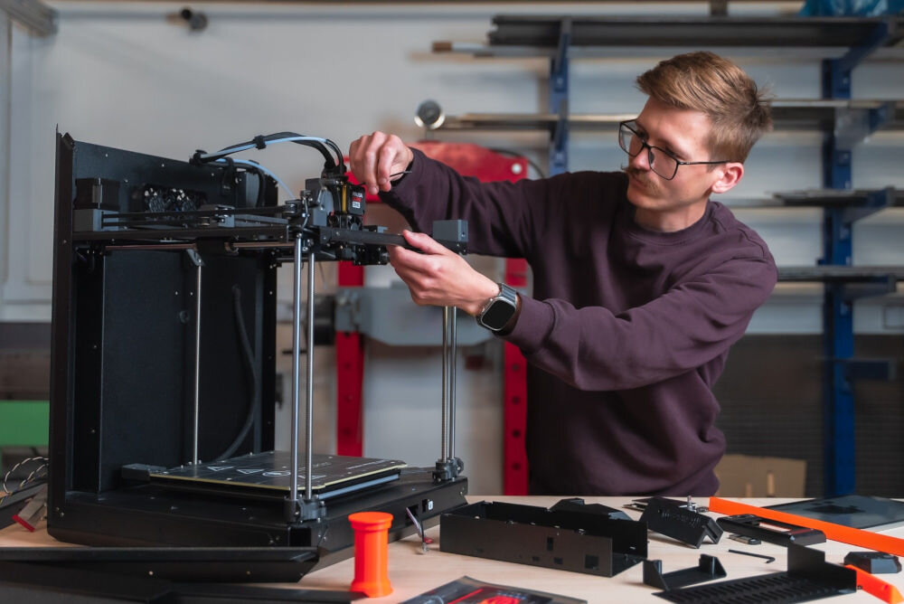 Drukarka 3D PRUSA Core One+ Wi-Fi, LAN + Zestaw do montażu Mężczyzna w okularach składający konstrukcję drukarki 3D przy stole warsztatowym filamenty Flex, łatwe ładowanie, elastyczne modele