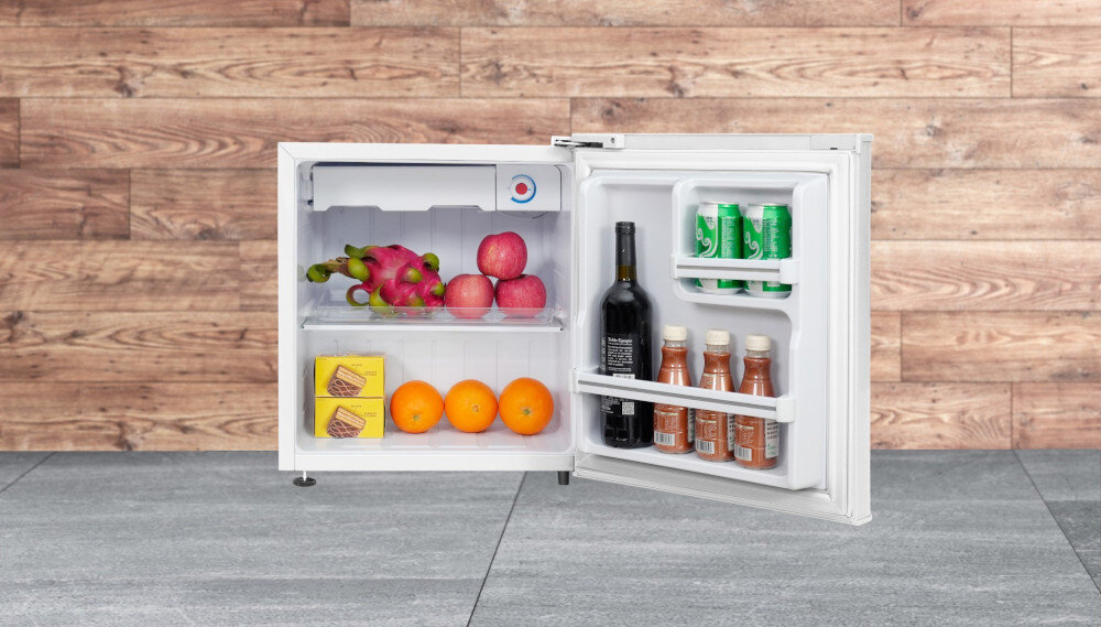 LODÓWKA TCL RF045DWE0 Na zdjęciu widać otwartą chłodziarkę ustawioną na szarej podłodze z płytkami, na tle ściany wykończonej panelami w kolorze jasnego drewna. W środku lodówki znajdują się smoczy owoc i jabłka na górnej półce, pomarańcze i dwa kartoniki na dolnej, a na drzwiach butelka wina, trzy butelki sosu oraz trzy zielone puszki napoju kompaktowa lodówka funkcjonalność higiena stabilność półka XL małe przestrzenie