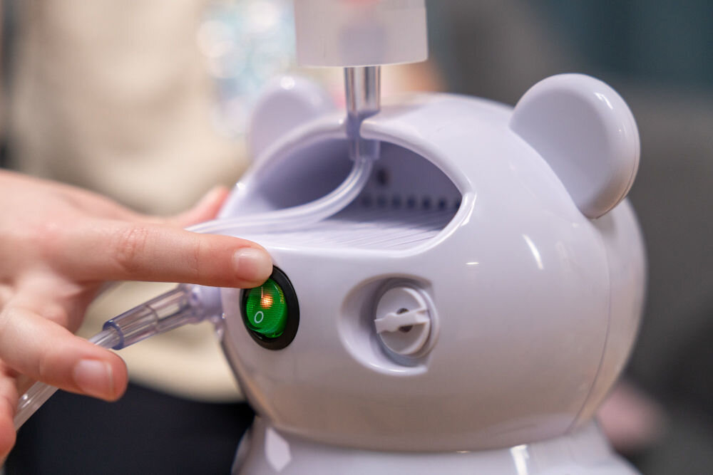 Biały inhalator w kształcie misia z widocznym przyciskiem z zielonym światłem i symbolem \'I/O\'. Osoba dotyka przycisku, a z urządzenia wychodzi przezroczysta rurka.