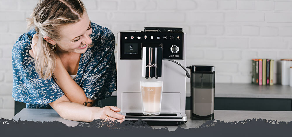 Ekspres do kawy z logo Melitta i LatteSelect, z filiżanką kawy na tacy. W tle widoczna ceglana ściana i kilka książek na blacie.