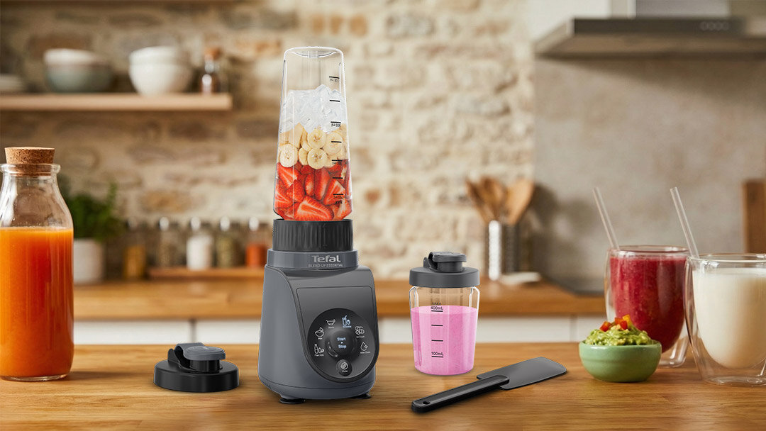 Na blacie kuchennym znajduje się blender z przezroczystym pojemnikiem wypełnionym pokrojonymi owocami. Obok blendera leży pokrywka, dodatkowy pojemnik z różowym płynem oraz szpatułka.