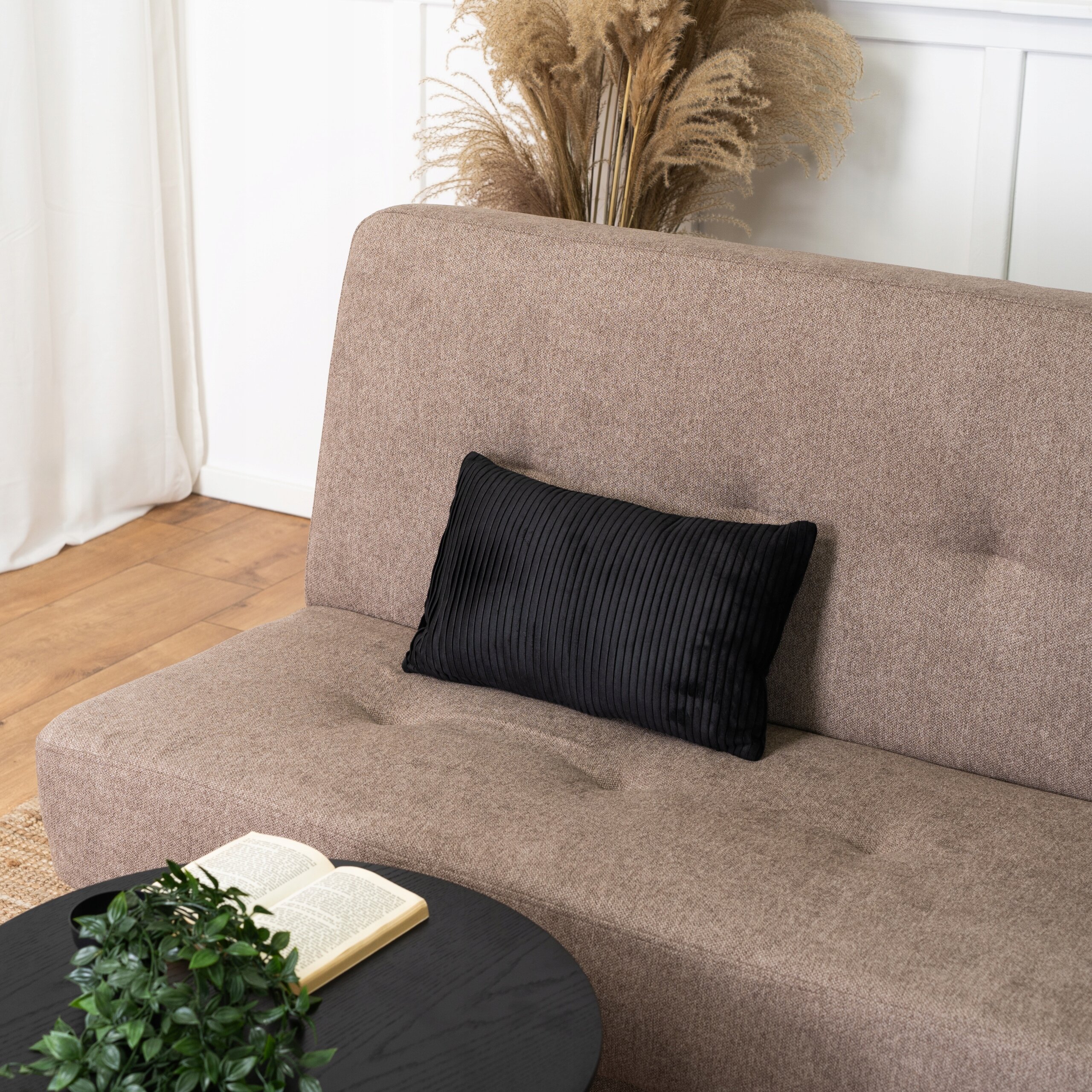 Brązowa sofa z czarną poduszką, obok której znajduje się otwarta książka na okrągłym, czarnym stoliku. W tle widoczne są suszone trawy dekoracyjne.