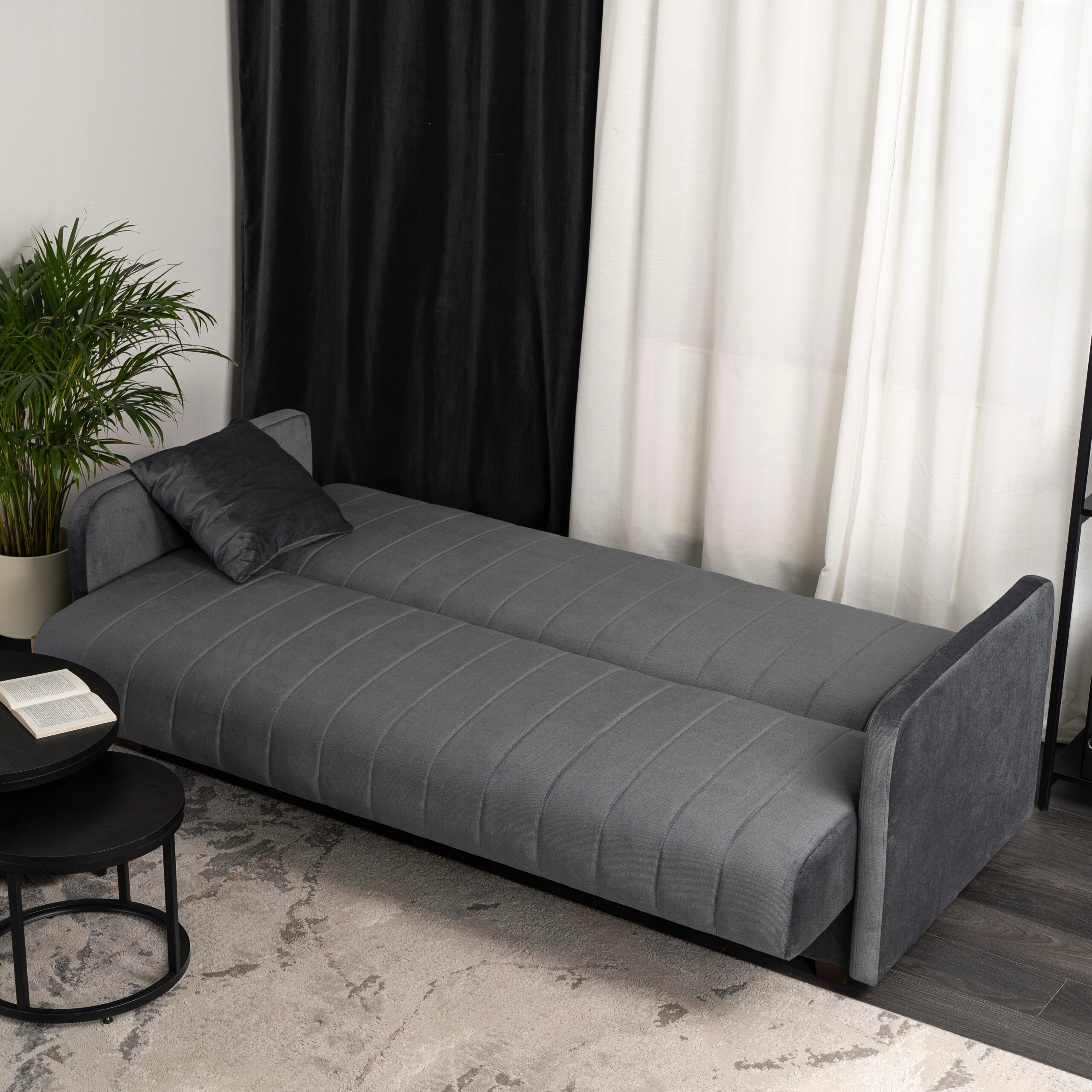 Szara, rozkładana sofa z funkcją spania, ustawiona w pomieszczeniu z ciemnymi i jasnymi zasłonami. Obok sofy znajduje się okrągły stolik z książką i rośliną w doniczce.