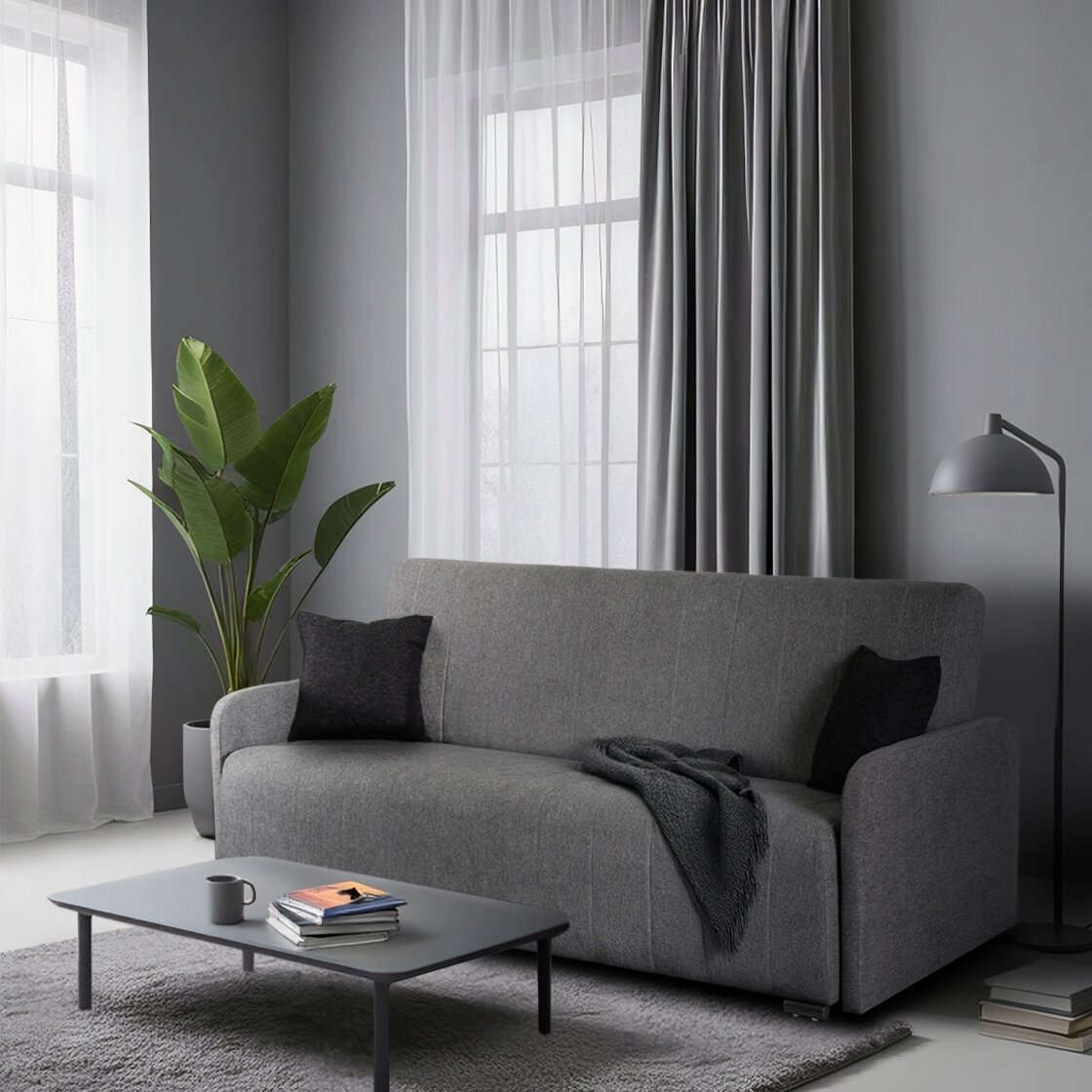Szara rozkładana sofa z funkcją spania, na której leży ciemny koc i dwie poduszki. Przed sofą znajduje się niski stolik z kubkiem i książkami, a obok stoi lampa podłogowa i roślina w doniczce.