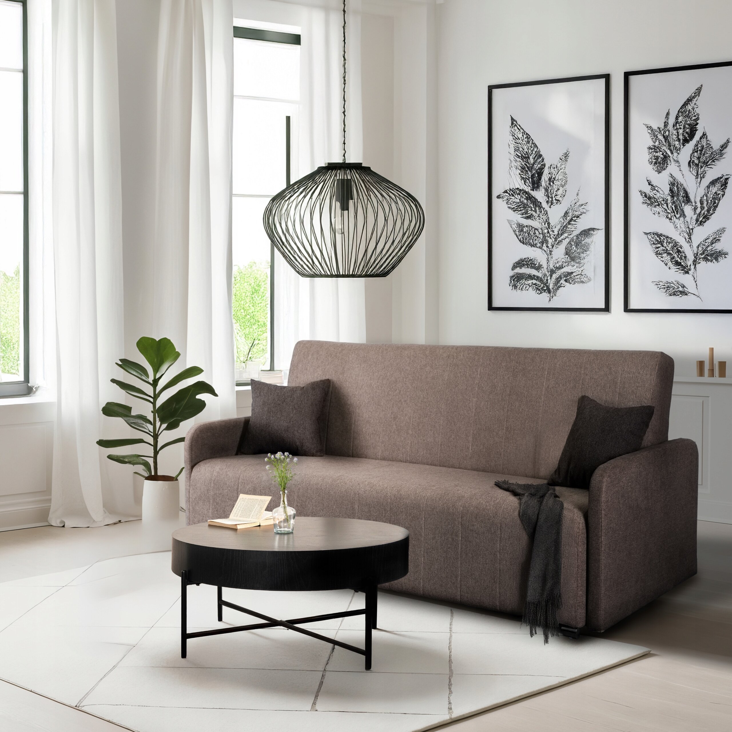 W pokoju znajduje się beżowa sofa z funkcją spania, przed którą stoi okrągły, czarny stolik z książką i wazonem z kwiatami. Na ścianie wiszą dwa obrazy z motywem roślinnym, a z sufitu zwisa nowoczesna lampa.