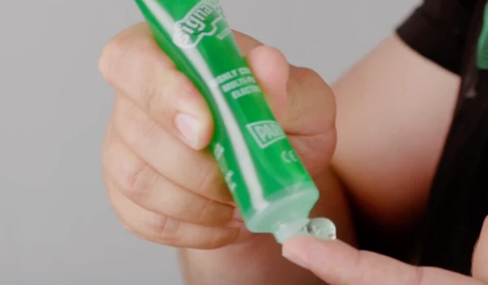 Osoba wyciska zielony żel z tubki na palec. Na tubce widoczny jest tekst: \'CONDUCTIVE GEL\'.