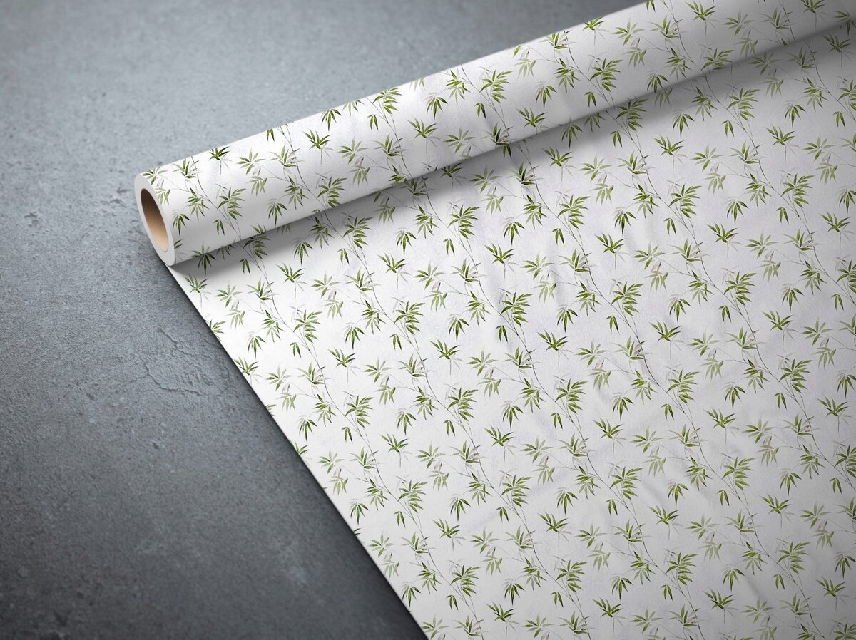 Na obrazie widoczna jest rozwinięta rolka ceraty z wzorem zielonych liści na białym tle. Cerata leży na szarej powierzchni.