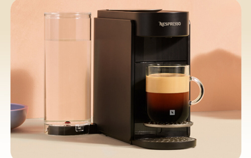 Czarny ekspres do kawy z logo \'Nespresso\' na górze, obok przezroczysty pojemnik na wodę. Na ekspresie stoi szklanka z kawą z pianką.