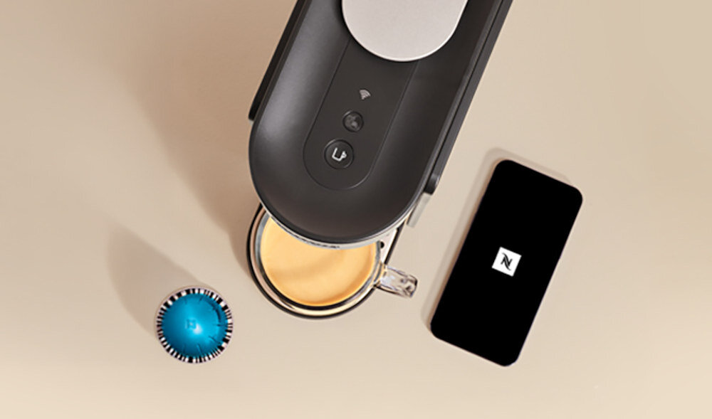 Na obrazie widoczny jest czarny ekspres do kawy z filiżanką pełną kawy oraz kapsułką w kolorze niebieskim. Obok znajduje się czarny smartfon z logo \'N\' na ekranie.