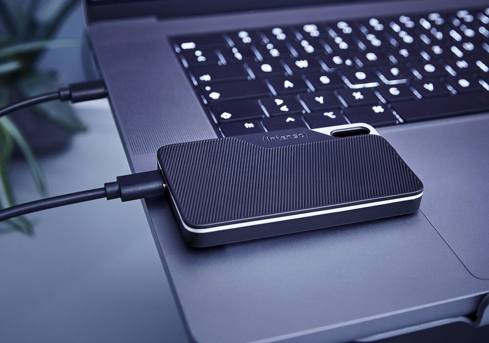 Dysk INTENSO TX100 Prostokątny dysk zewnętrzny o prążkowanej fakturze podłączony kablem do laptopa i leżący na jego obudowie, nośnik, pojemność, dioda LED