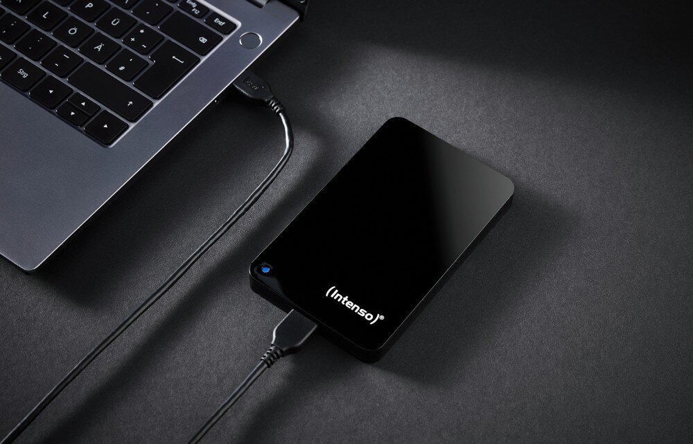 Dysk INTENSO Memory Drive Przenośny dysk twardy z zaświeconą niebieską diodą leży na ciemnym blacie, podłączony kablem USB do widocznego fragmentu laptopa, pojemność, prędkość, interfejs, kompatybilność