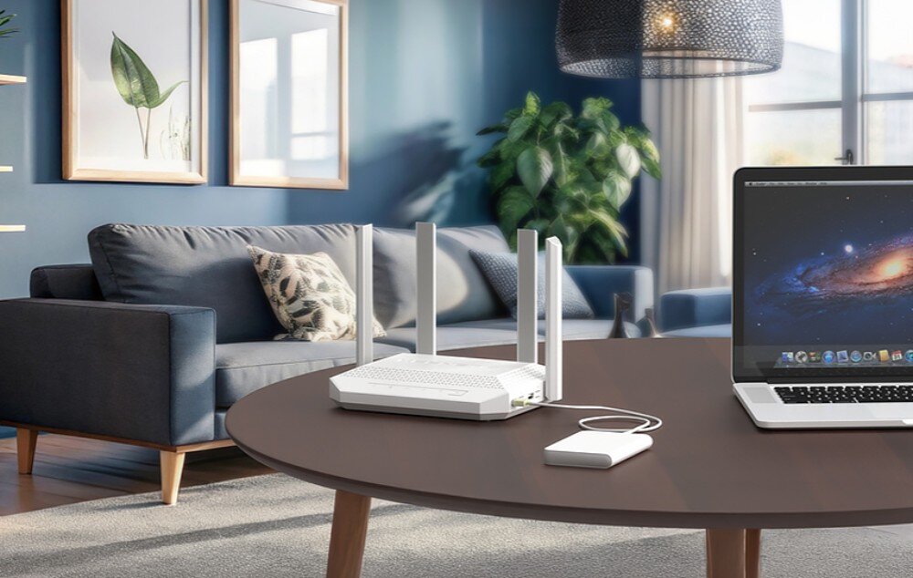 Router KEENETIC Hero KN-1012 Router z czterema antenami stoi na ciemnym, drewnianym stole obok laptopa i małego dysku zewnętrznego w jasnym salonie z niebieską ścianą, szarą sofą i rośliną donickową