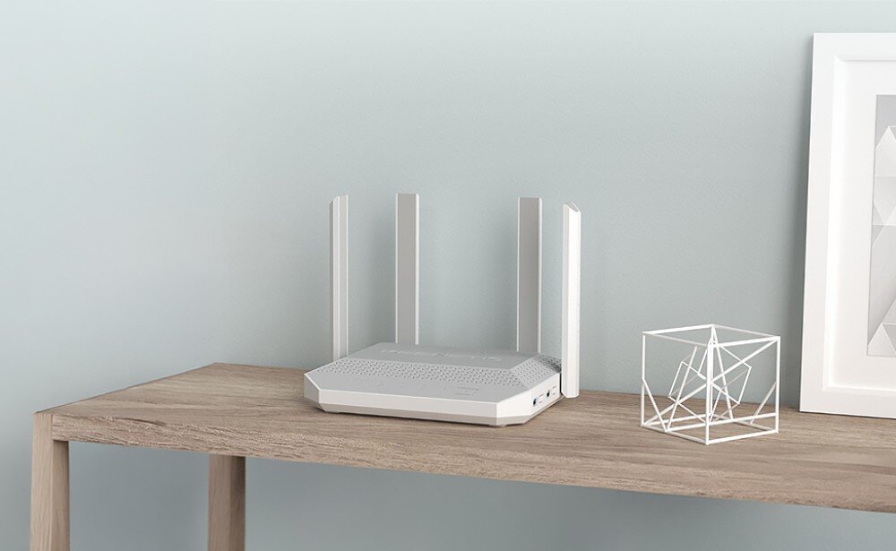 Router KEENETIC Hero KN-1012 Biały router z czterema antenami stoi na drewnianym blacie obok białej ramki na zdjęcia i geometrycznej dekoracji na tle jasnej ściany