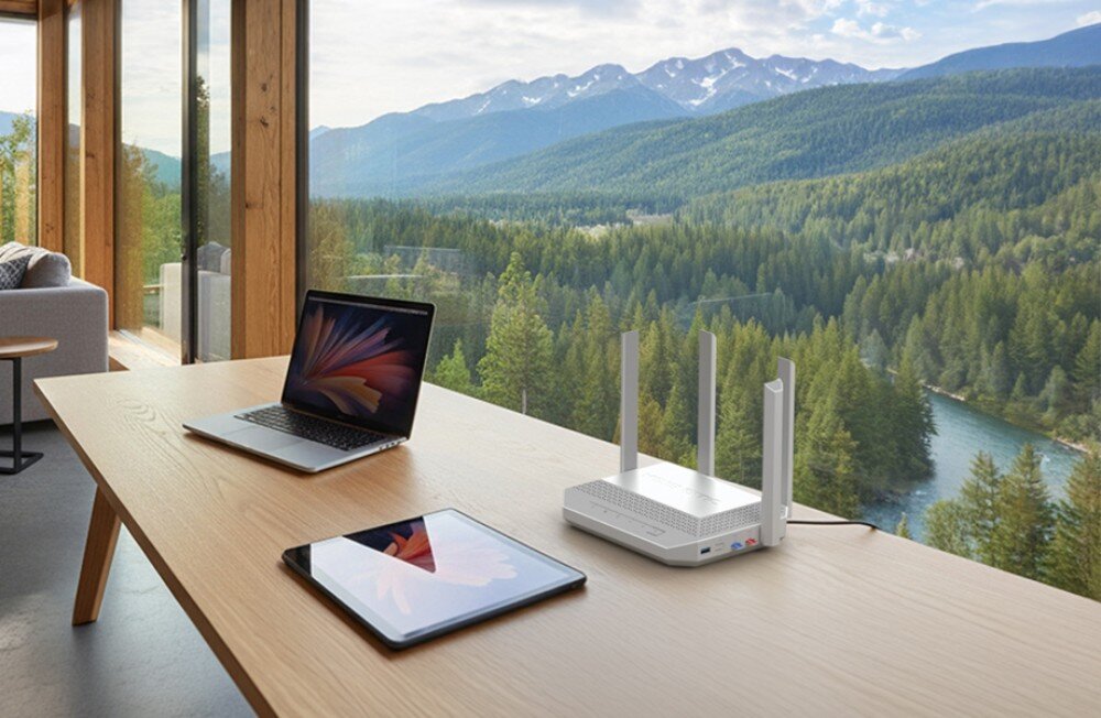 Router KEENETIC Hopper 4G+ Biały router z czterema antenami stoi na jasnym, drewnianym stole obok otwartego laptopa i tabletu, a w tle przez duże okno widać górski krajobraz i las