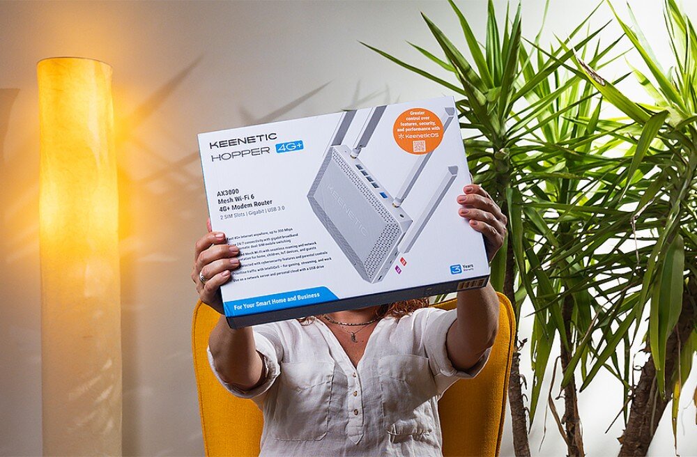 Router KEENETIC Hopper 4G+ Osoba w białej koszuli trzyma przed twarzą duże białe pudełko z grafiką i opisem technicznym routera, siedząc w żółtym fotelu obok wysokiej rośliny