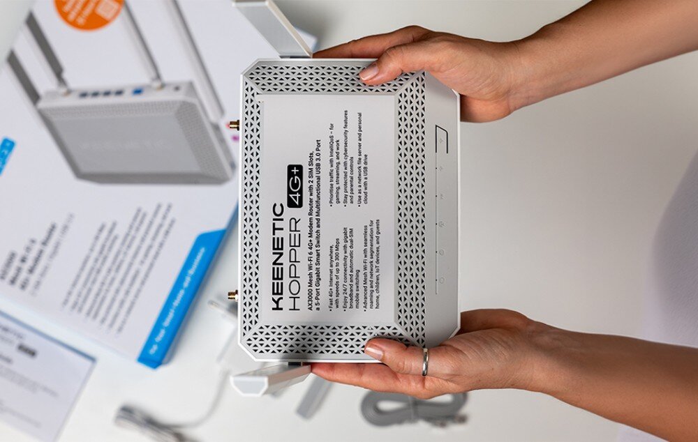 Router KEENETIC Hopper 4G+ Widok z góry na dłonie trzymające biały router, na którego obudowie naklejona jest etykieta z parametrami technicznymi, obok leży opakowanie i akcesoria