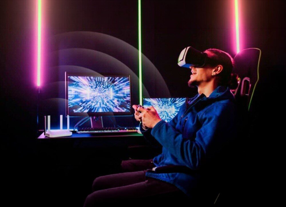 Router KEENETIC Titan 3 postać w goglach do wirtualnej rzeczywistości siedzi w fotelu przed monitorem, a obok na biurku znajduje się białe urządzenie z antenami w ciemnym pokoju z neonowym oświetleniem