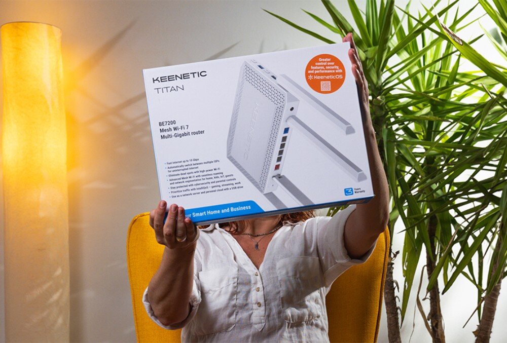 Router KEENETIC Titan 3 postać w białej koszuli siedzi w żółtym fotelu i trzyma przed sobą pudełko z ilustracją urządzenia sieciowego na tle zielonej rośliny