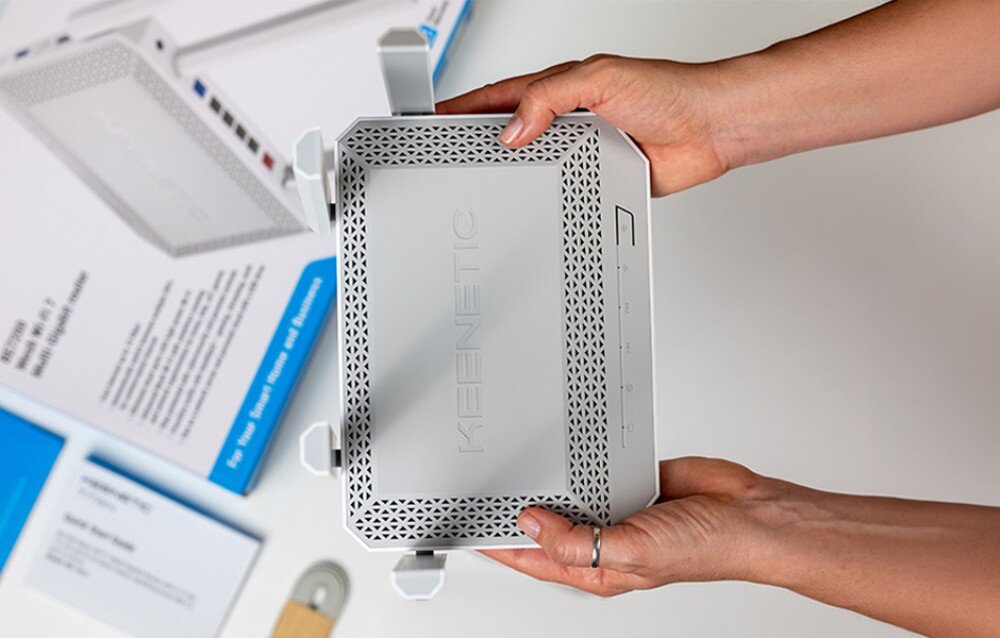 Router KEENETIC Titan 3 dłonie trzymają białe urządzenie z otworami w obudowie nad białym blatem, na którym leżą dokumenty i fragment opakowania
