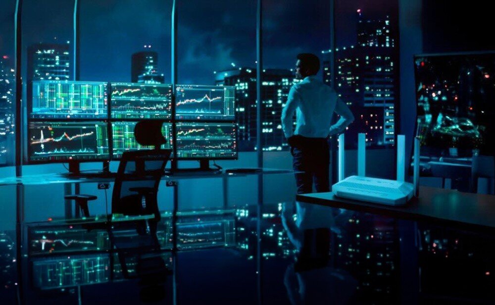 Router KEENETIC Titan 3 postać w biurze patrzy przez okno na światła miasta nocą obok ekranów z wykresami, a na blacie z przodu stoi białe urządzenie z antenami