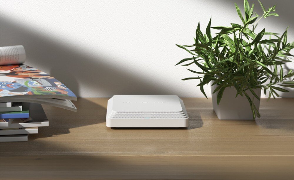 Punkt dostępu KEENETIC Orbiter Pro AC1300 Białe płaskie urządzenie leżące na drewnianym blacie obok rośliny doniczkowej i stosu magazynów