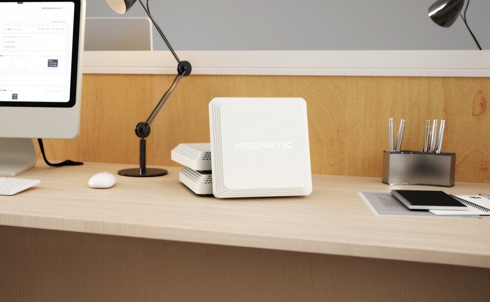 Punkt dostępu KEENETIC Orbiter Pro AC1300 Białe urządzenie ustawione pionowo na jasnym biurku w sąsiedztwie monitora i akcesoriów biurowych
