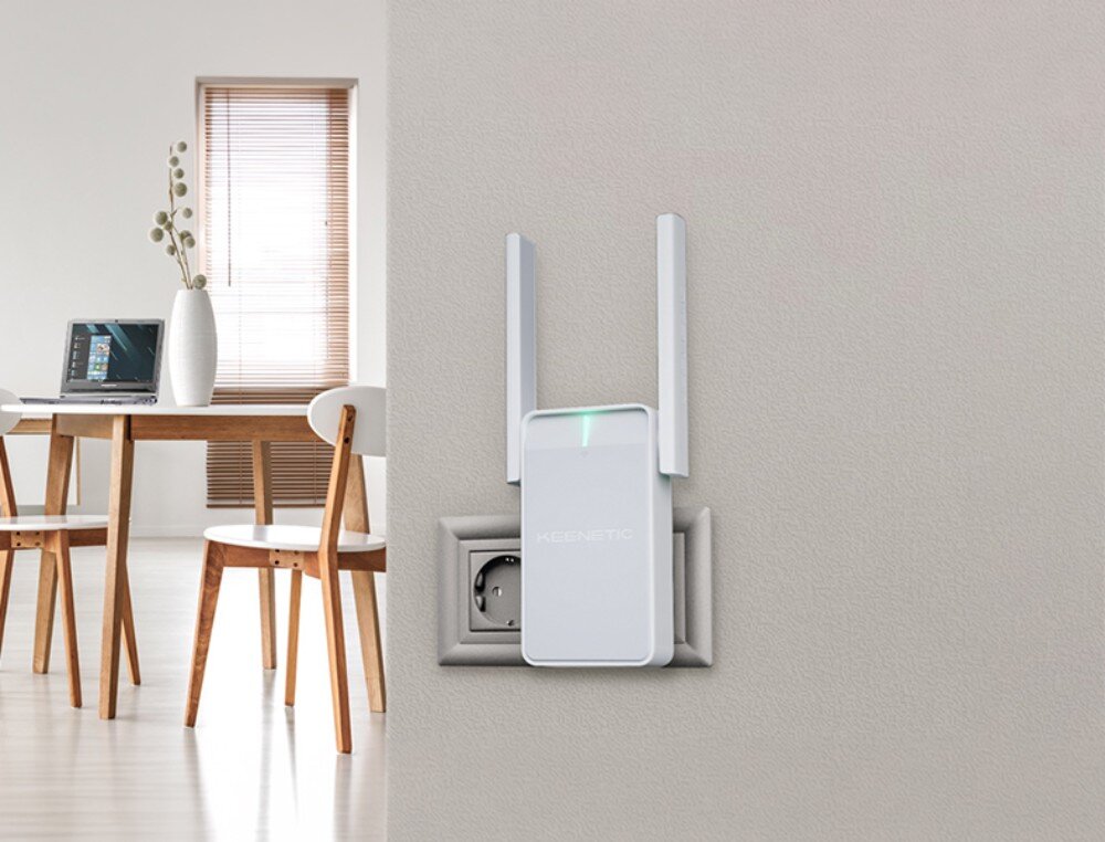 Wzmacniacz sieci KEENETIC Buddy 4 Jasne urządzenie sieciowe z dwiema pionowymi antenami wpięte do gniazdka na szarej ścianie, w tle widać fragment jadalni z laptopem na stole