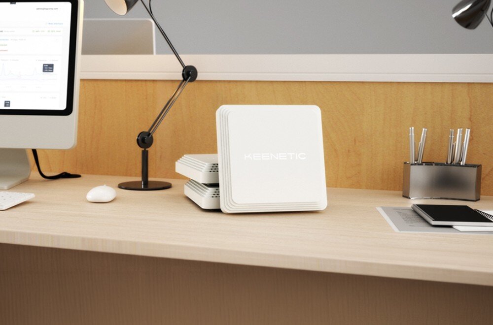 Punkt dostępu KEENETIC Voyager Pro Białe kwadratowe urządzenie z napisem 'KEENETIC' ustawione pionowo na jasnym biurku obok monitora i lampy