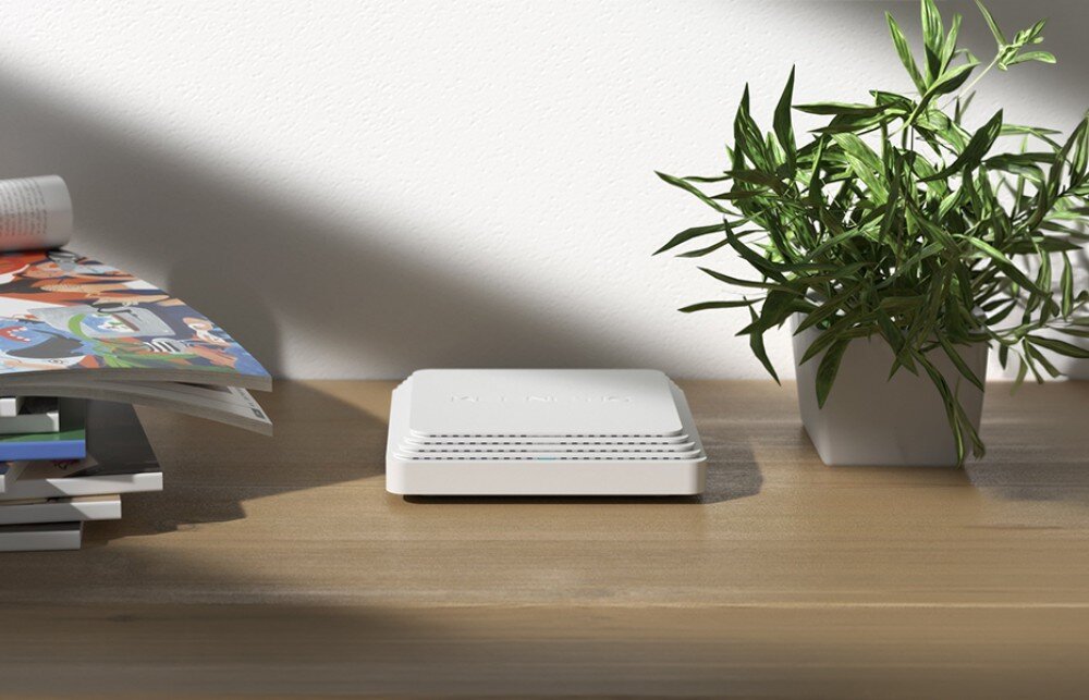 Punkt dostępu KEENETIC Voyager Pro Białe kwadratowe urządzenie leżące płasko na drewnianym blacie obok zielonej rośliny i stosu czasopism