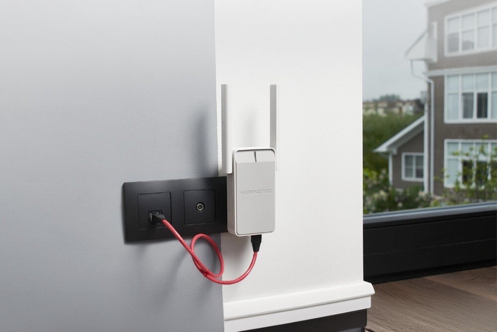 Wzmacniacz sieci KEENETIC Buddy 5 białe urządzenie z dwiema antenami wpięte do gniazdka na białej ścianie i połączone czerwonym kablem z gniazdem sieciowym obok okna