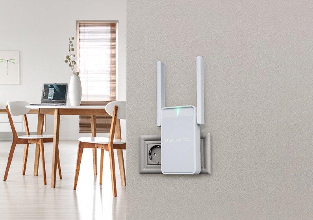 Wzmacniacz sieci KEENETIC Buddy 5 białe urządzenie z dwiema antenami wpięte do gniazdka na szarej ścianie, w tle stół z krzesłami i laptopem w jasnym pokoju