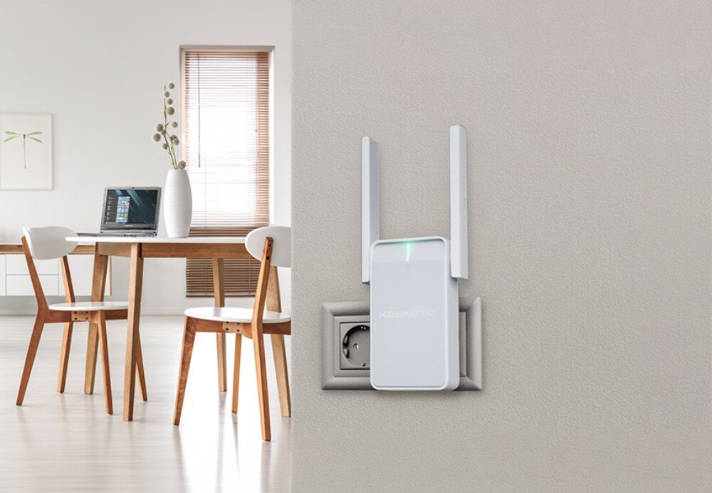Wzmacniacz sieci KEENETIC Buddy 6 Białe urządzenie sieciowe z dwiema pionowymi antenami wpięte do kontaktu na jasnej ścianie, w tle stół z laptopem