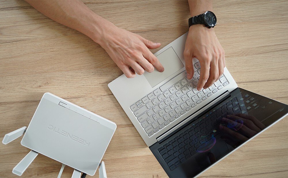 Router KEENETIC Speedster Widok z góry na dłonie piszące na białym laptopie na drewnianym blacie obok białego rutera