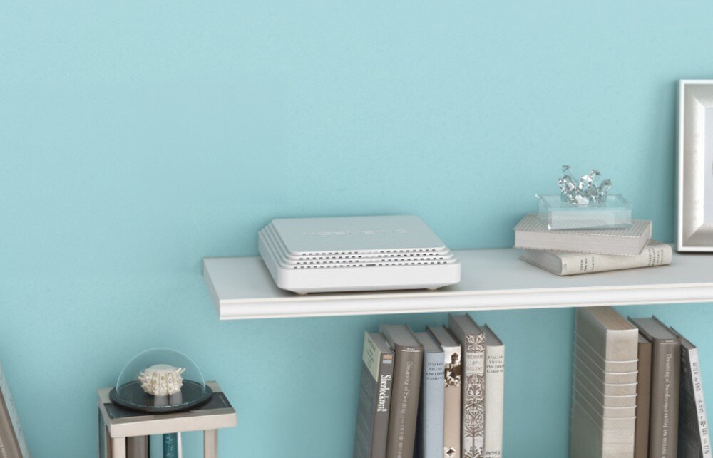 Router KEENETIC Sprinter SE białe urządzenie o kwadratowej formie leży na białej półce obok rzędu książek i ozdobnych przedmiotów na tle jasnej błękitnej ściany