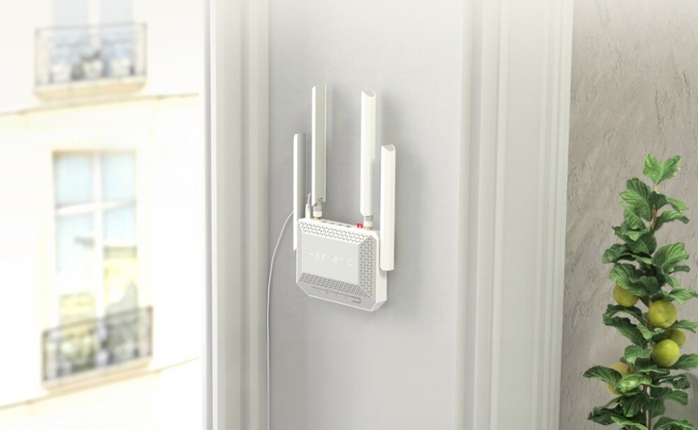 Router KEENETIC Speedster 4G+ Biały ruter zamontowany pionowo na jasnej ścianie obok okna z widokiem na budynki i fragmentu rośliny